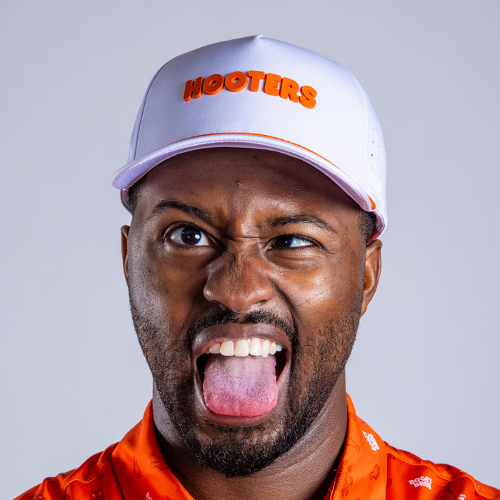 A man in an orange shirt makes a silly face while wearing the Hooters - Performance Golf Rope Hat - Snapback, which is sweat stain resistant. The white snapback stands out against the plain, light gray background.