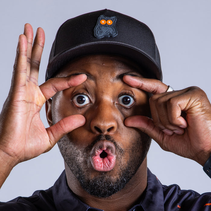 A man in the Night Owl Performance Golf Hat Snapback, featuring an owl patch and sweat stain resistant material, opens his eyes wide with his fingers and makes an O with his mouth against a plain light gray background.