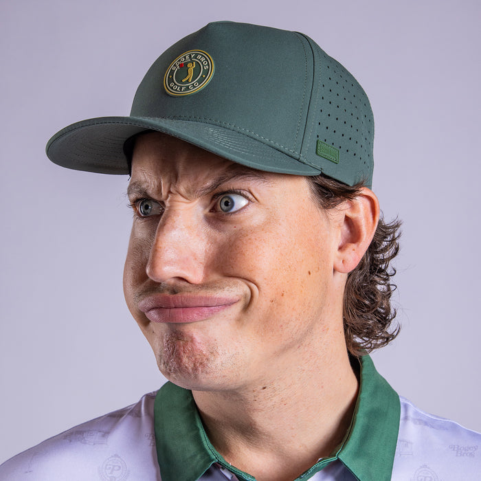 A man with blue eyes, light skin, and wavy brown hair wears the Peasants Stamp - Performance Golf Hat - Snapback (green, round logo) and a collared shirt. He makes a comical pouty face against a plain, light background.