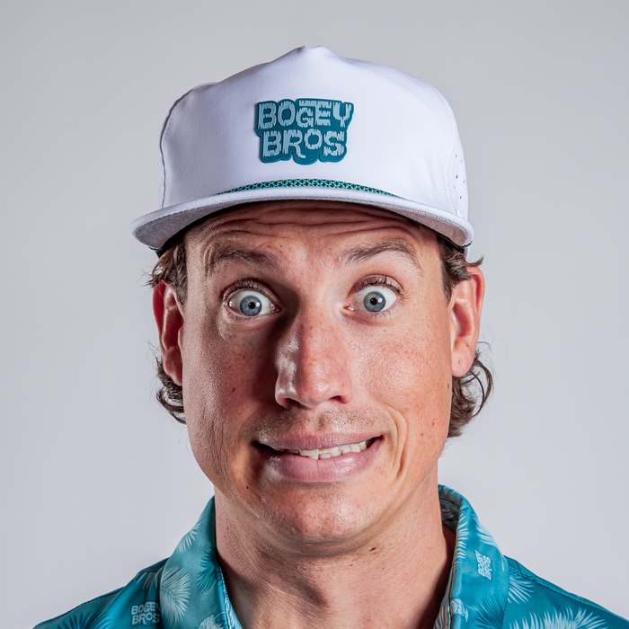 A man with wide blue eyes and a nervous, toothy smile wears the Tiki Flat Bill Unstructured Flat Bill Hat - Snapback and a teal patterned shirt. He has light skin, brown hair showing under the hat, and stands against a plain light gray background.