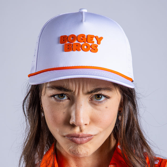 A woman with long brown hair wears the Wingin' It - Performance Golf Rope Hat - Snapback, featuring bold orange "BOGEY BROS" text and an orange stripe. She pairs it with an orange patterned shirt against a plain light gray background.