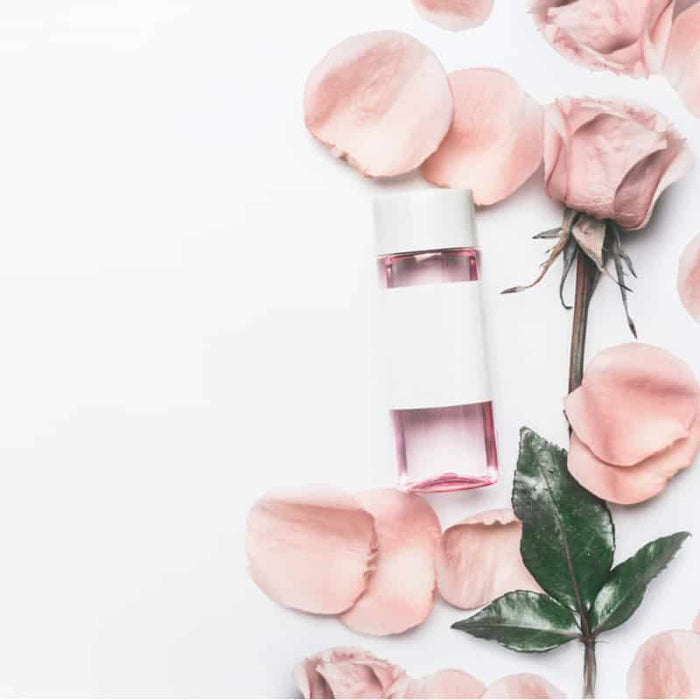 A clear bottle of Anti-Aging Face & Body Oil | Privai surrounded by soft pink rose petals and green leaves, conveying a natural skincare and body care theme.