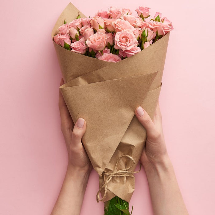 Hands holding a bouquet of pink roses wrapped in brown paper on a soft pink background, evoking a fresh and natural aesthetic for Anti-Aging Face & Body Oil | Privai skincare.