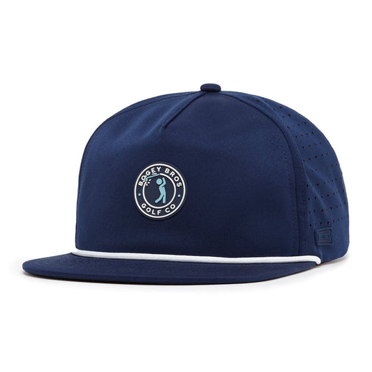 The Bogeyman Stamp Navy Unstructured Flat Bill Hat is a navy snapback featuring white accent piping, an embroidered circular “Bogey Bros Golf Co” golfer logo, side ventilation holes, and performance material construction.