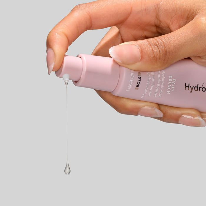 Hand holding a pink bottle dispensing a drop of moisturizing serum from Daily Drench | HydroPeptide skincare product against a plain gray background