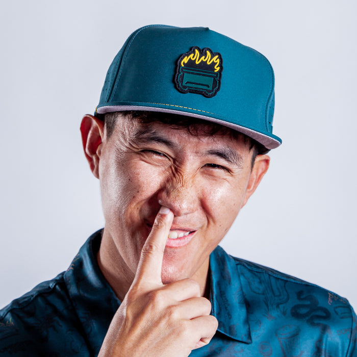 Smiling and squinting playfully, a person in a patterned teal shirt picks their nose while wearing the Dumpster Fire - Unstructured Flat Bill Hat - Snapback against a plain light background.