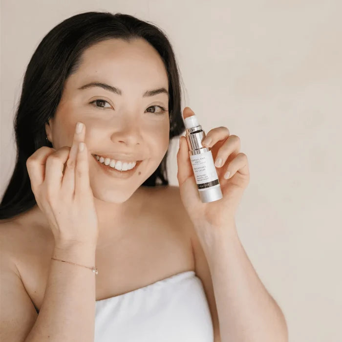 Smiling woman with clear skin holding a bottle of Enlightened Eye Cream | DermaplanePro while applying the product near her eye in a skincare routine setting.