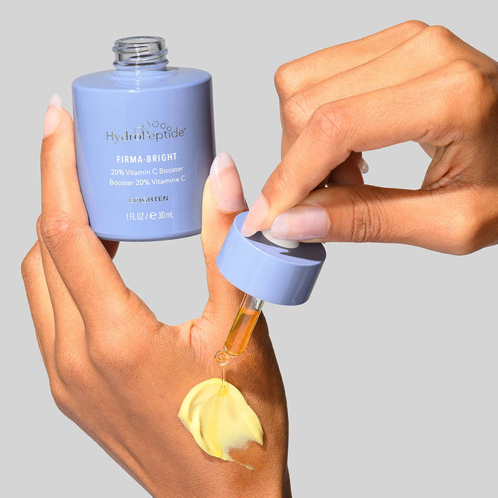 Hand applying yellow cream with a dropper from a light purple bottle of Firma-Bright | HydroPeptide vitamin C skincare booster against a neutral background.