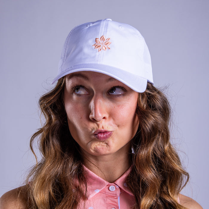 A woman in a white "In Bloom - Dad Hat" with a peach flower emblem and adjustable strap, wearing a pink polka dot shirt, makes a kissing face while looking to her left with wide eyes.