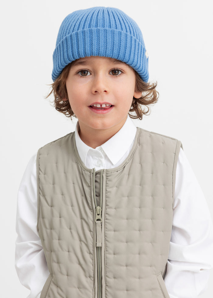 Child wearing a green quilted vest and a blue knitted hat.