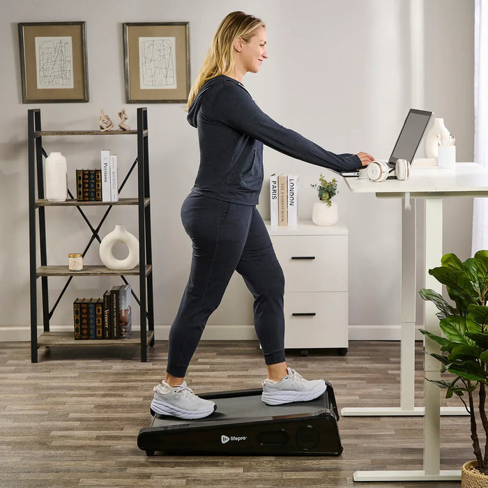 Woman walking on the PacerMini treadmill in front of a standing desk while typing on her laptop, headphones resting beside it.