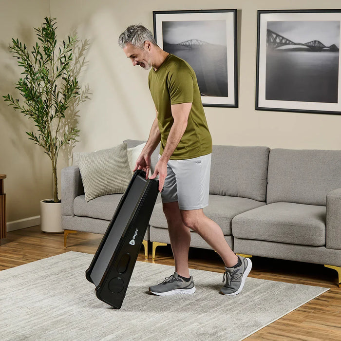Man lifting the folded PacerMini treadmill by its edge to carry it across a living room floor.