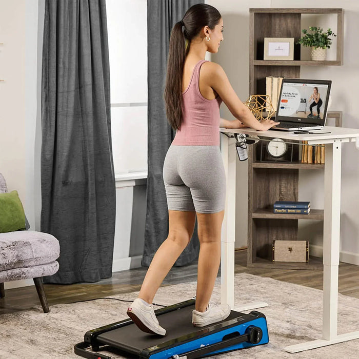 Woman seen from behind walking on the PacerMini treadmill in a home office, resting her hand on a standing desk with a monitor.