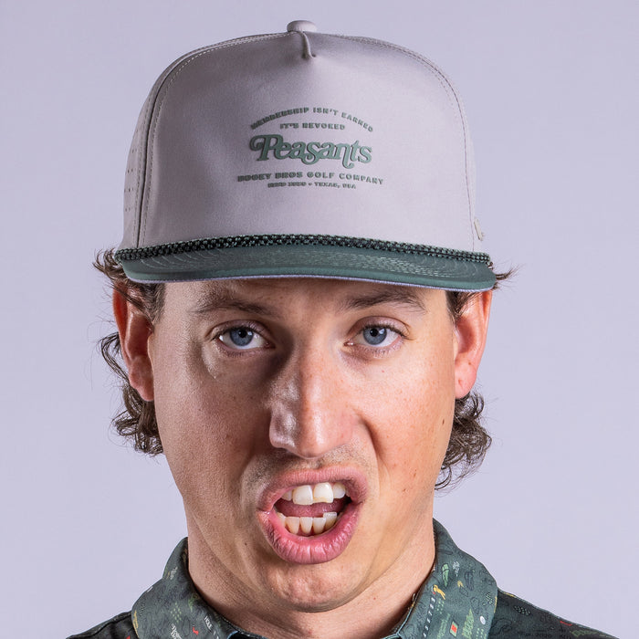 A person faces the camera wearing the Revoked - Unstructured Flat Bill Hat - Snapback in light gray and dark green. Their mouth is open mid-expression, revealing upper teeth, brown hair peeks from under the cap, and the plain background is light.