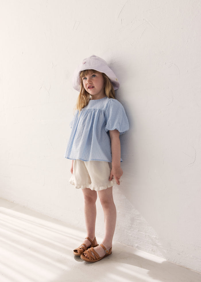Pige iført kortærmet bluse til børn med puffærmer og fint broderimønster, lyse shorts og solhat

Girl wearing a short-sleeved blouse with puffed sleeves and a fine embroidery pattern, light shorts and a sun hat