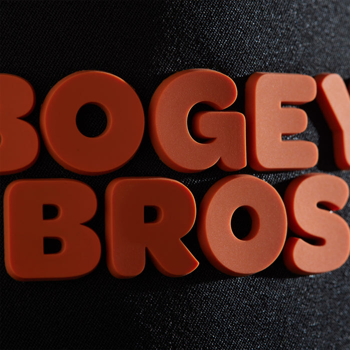 Bold, rounded red-orange letters spelling "BOGEY BROS" appear in two lines on a textured black background. Inspired by the Wingin' It Unstructured Flat Bill Hat - Snapback, the letters cast subtle shadows for a 3D look.