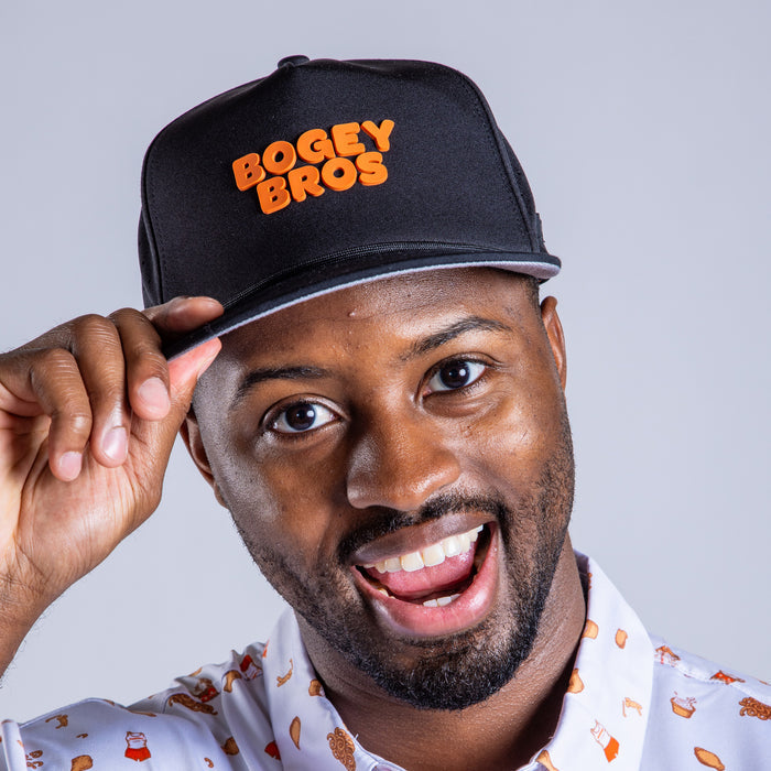 A smiling man tilts his Wingin' It - Unstructured Flat Bill Hat - Snapback. He wears a white shirt with cartoon hamburgers and fries on it, standing against a plain light gray background.